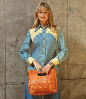 A woman in a denim dress with yellow accents stands against a textured wall, holding the Bed|Stü Orchid handwoven leather bag featuring an antique metal buckle in front of her, effortlessly embodying modern timeless design.