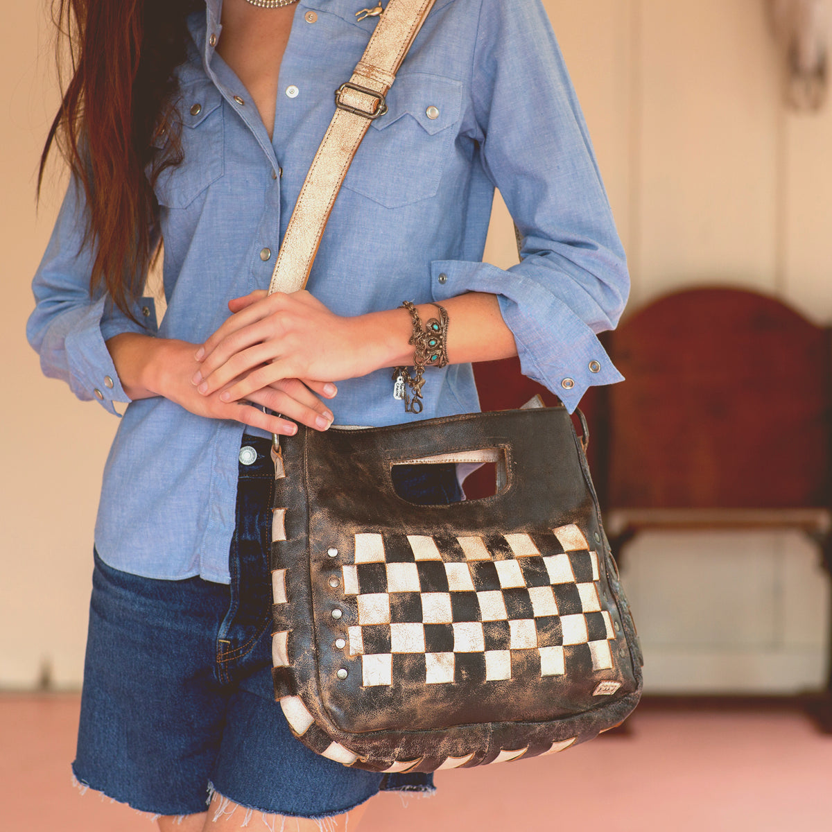 Wearing a blue button-up shirt and denim shorts, a person holds the Bed|Stü Orchid shoulder bag—a black and white checkered accessory featuring a modern, timeless design with an antique metal buckle.