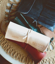 The Bed|Stü Prepped Leather Wrap, an elegant cream leather roll-up organizer embossed with the brand logo, rests atop a red leather pouch and other leather goods.