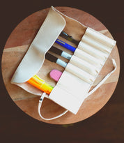 The Bed|Stü Prepped Leather Wrap—a white organizer designed to hold several pens, a yellow highlighter, and a pink eraser—is stylishly displayed on a round wooden surface, making it the perfect storage solution for artists who value both organization and elegance.