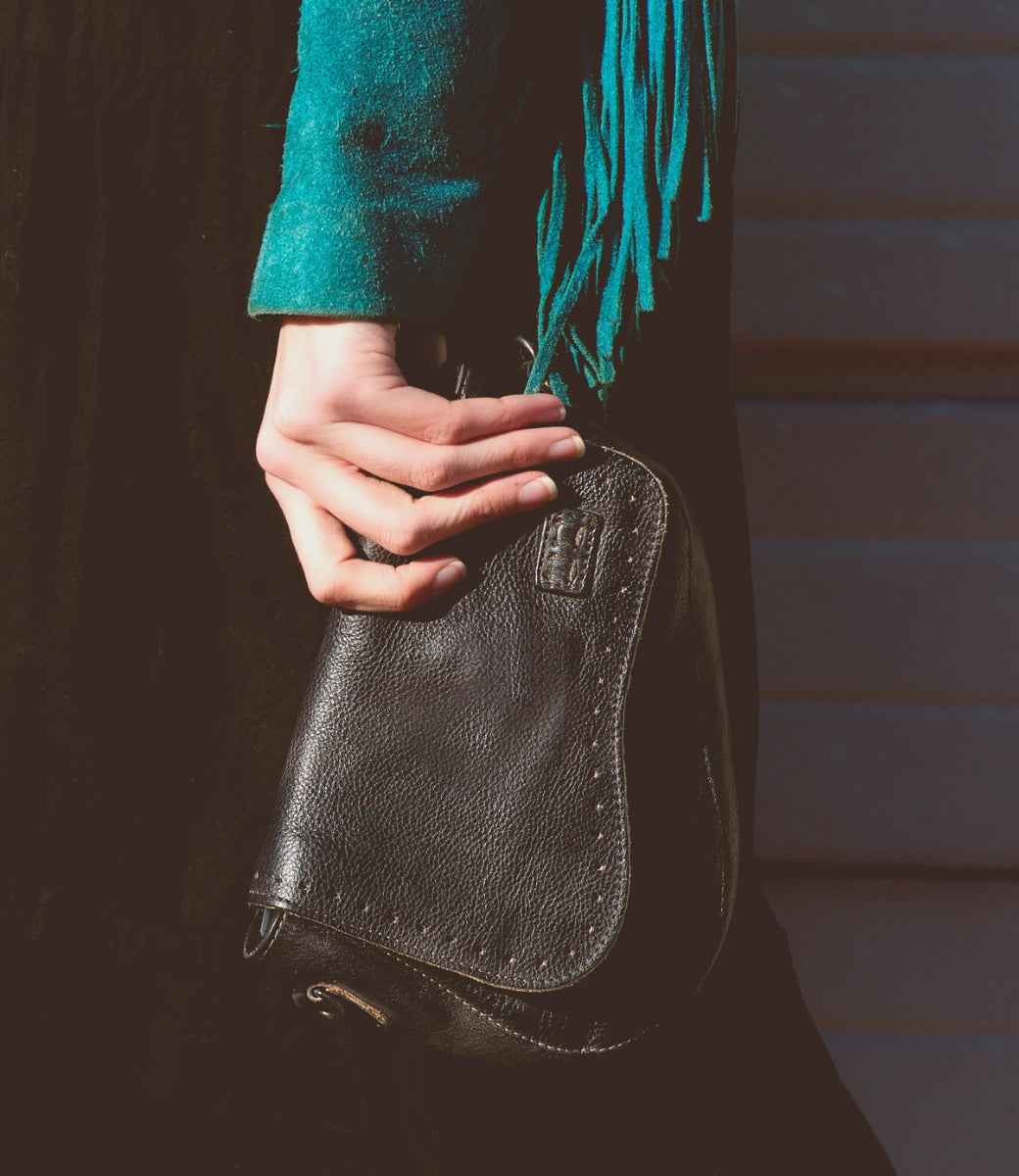 A person wearing a teal sleeve holds the Bed|Stü Priscilla small black leather crossbody bag against a dark background.