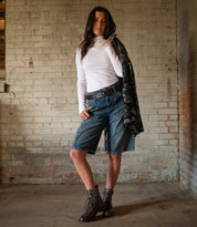 A woman stands indoors against a worn brick wall, wearing a white long-sleeve shirt, knee-length belted denim shorts paired with the Bed|Stü Proem—an artisanally detailed handwoven leather belt—and lace-up boots, while holding a patterned jacket over her shoulder.