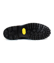 The image shows the black rubber treaded sole of the durable Bed|Stü Protege Trek boot, featuring a yellow hexagonal Vibram logo at the center.