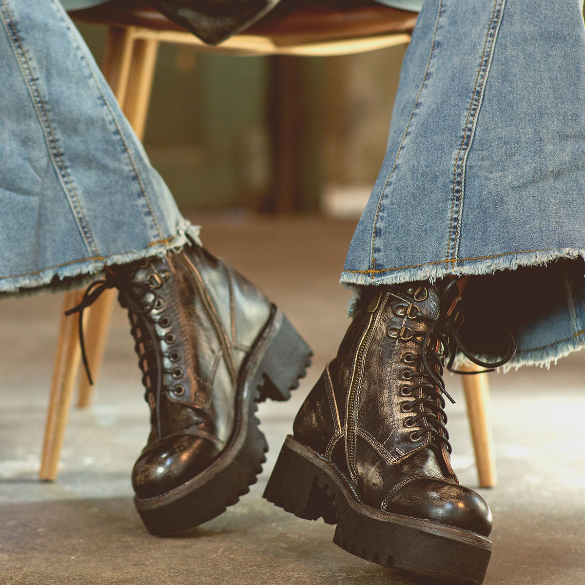 A person wearing distressed blue flared jeans and Bed|Stü Prudence Hi black boots featuring a chunky platform outsole, seated on a wooden chair with legs crossed at the ankles.