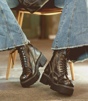 A person wearing distressed blue flared jeans and Bed|Stü Prudence Hi black boots featuring a chunky platform outsole, seated on a wooden chair with legs crossed at the ankles.