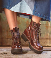 A person wearing the Bed|Stü Prudence Hi brown lace-up leather combat boots with side zippers and a chunky platform outsole stands on a wooden surface, dressed in a blue denim skirt.