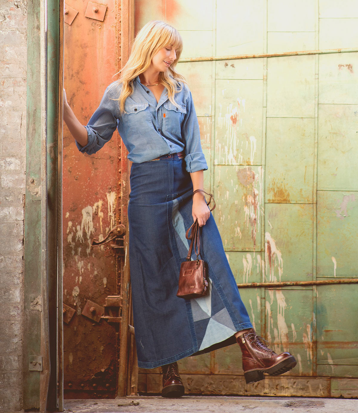 A woman in a denim shirt and patchwork denim skirt stands by a worn metal door, holding a small brown bag and wearing Bed|Stü Prudence Hi boots with a chunky platform outsole.