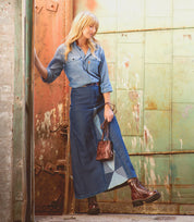 A woman in a denim shirt and patchwork denim skirt stands by a worn metal door, holding a small brown bag and wearing Bed|Stü Prudence Hi boots with a chunky platform outsole.