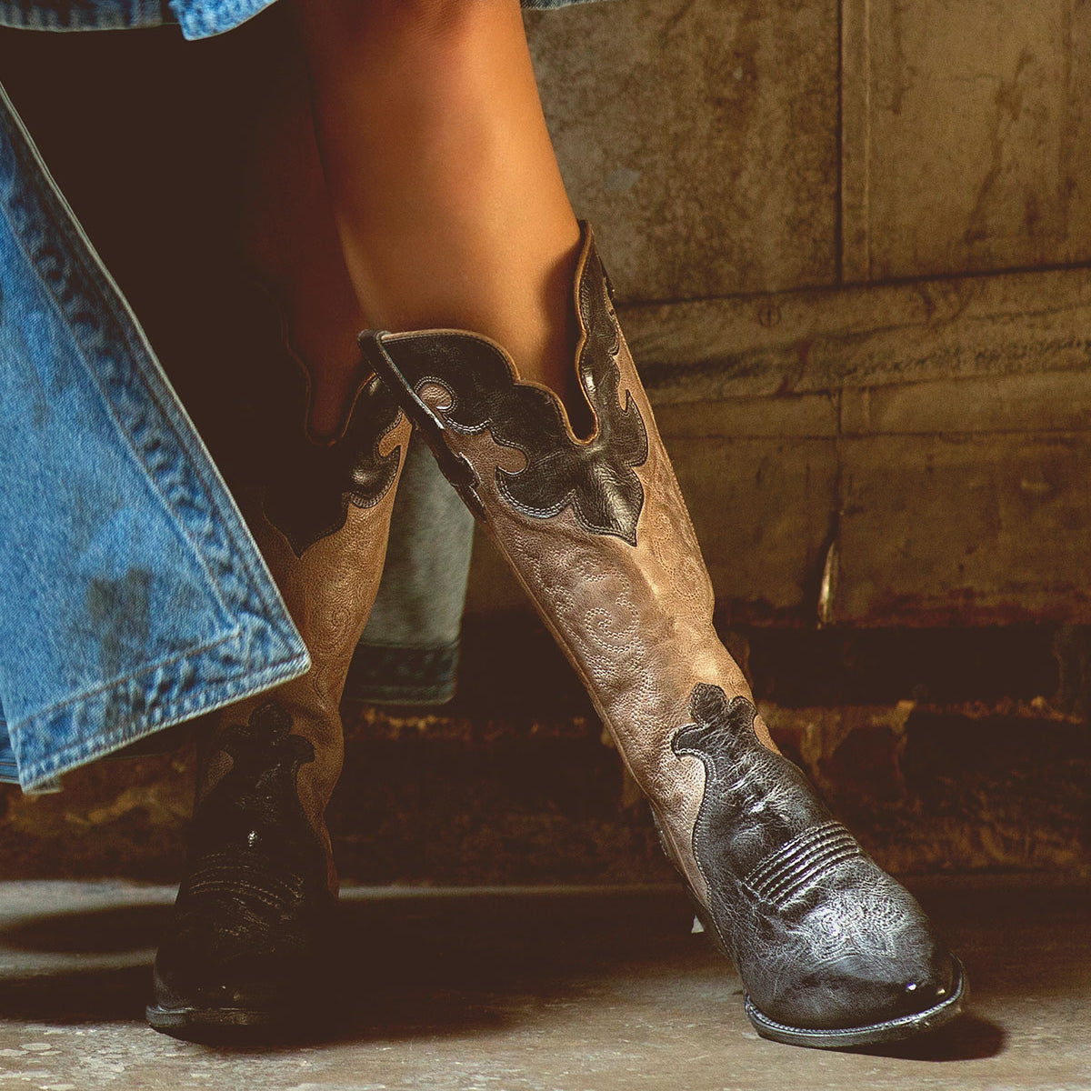 A person stands indoors on a concrete floor near a textured wall, wearing a denim skirt and Bed|Stü’s Queen brown Western boots featuring subtle Fleur-de-lis details.