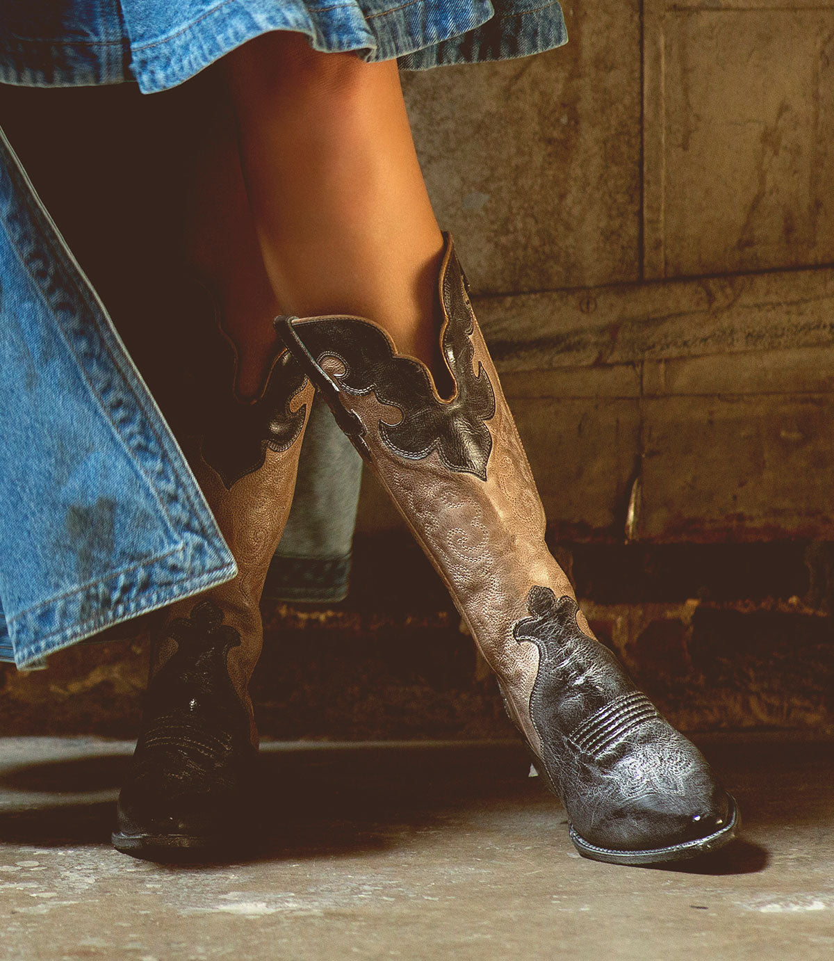 A person stands indoors on a concrete floor near a textured wall, wearing a denim skirt and Bed|Stü’s Queen brown Western boots featuring subtle Fleur-de-lis details.