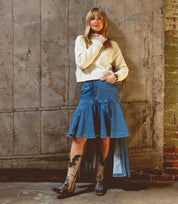 A woman stands against a worn industrial wall, wearing a cream sweater, a blue denim skirt with a flared hem, and the Bed|Stü Queen boots—black Western footwear crafted from vegetable-tanned leather with delicate fleur-de-lis embroidery along the shaft.
