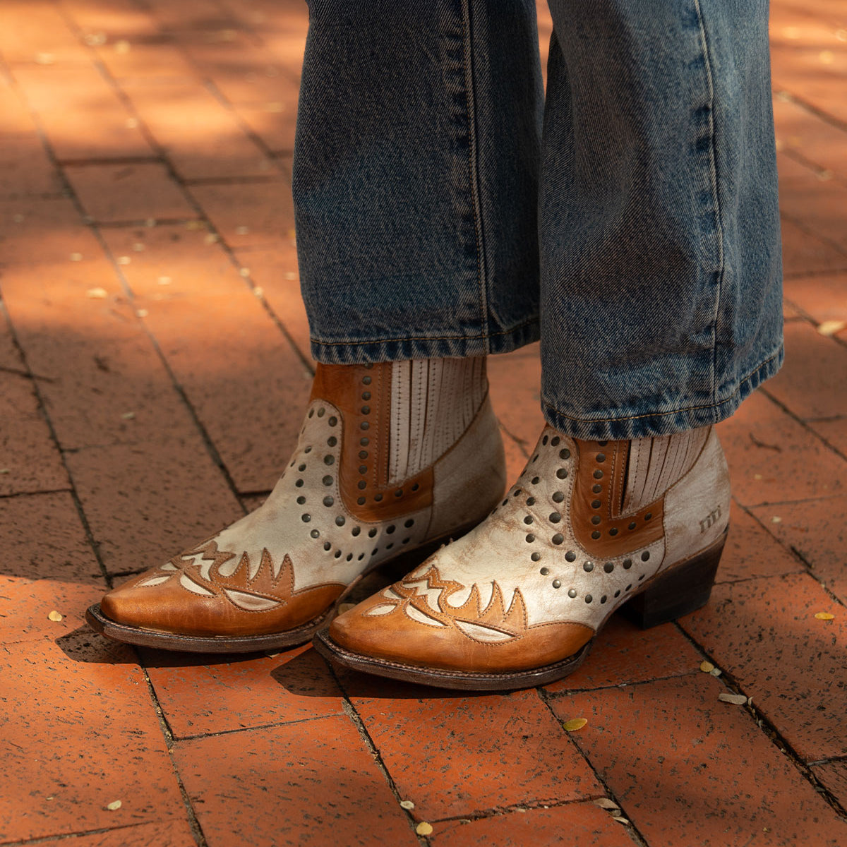 A person wearing the Bed|Stü Raise two-tone leather Western short boots with decorative stitching and studded accents, paired with blue jeans, standing on a red brick pavement.