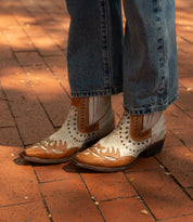 A person wearing the Bed|Stü Raise two-tone leather Western short boots with decorative stitching and studded accents, paired with blue jeans, standing on a red brick pavement.