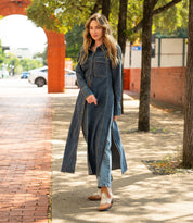 A woman stands on a sunny sidewalk wearing a long denim coat over jeans and tan Bed|Stü Raise Western short boots with a snip toe. Trees line the path, and red brick structures are in the background.