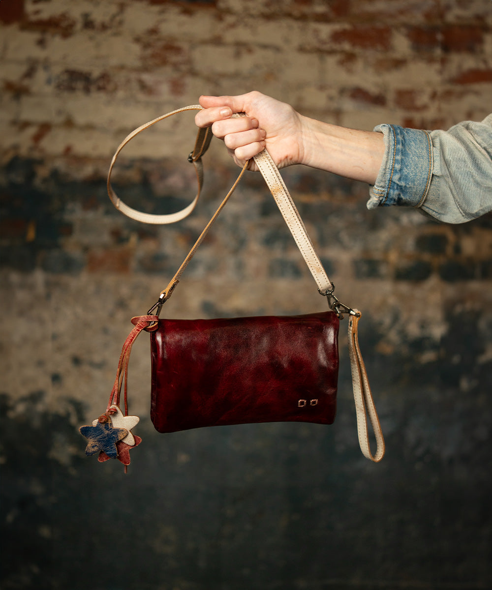 Hand holding a small burgundy leather crossbody bag with cream straps and a red, white, and blue star charm against a textured brick wall.