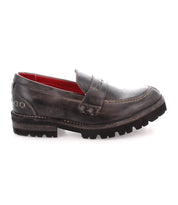 Presenting the Reina III by Bed|Stü—a single hand-finished dark brown leather penny loafer, featuring visible stitching, a rugged black sole, and striking red interior lining. Shown in side profile, this modern essential perfectly blends classic style with enduring craftsmanship.