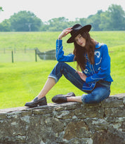 A woman in a blue embroidered shirt, rolled-up jeans, and a black hat sits on a stone wall in a grassy field, resting one hand on her hat. On her feet, she wears the Reina III penny loafers from Bed|Stü, crafted from hand-finished leather—a true modern essential that completes her effortlessly chic look.