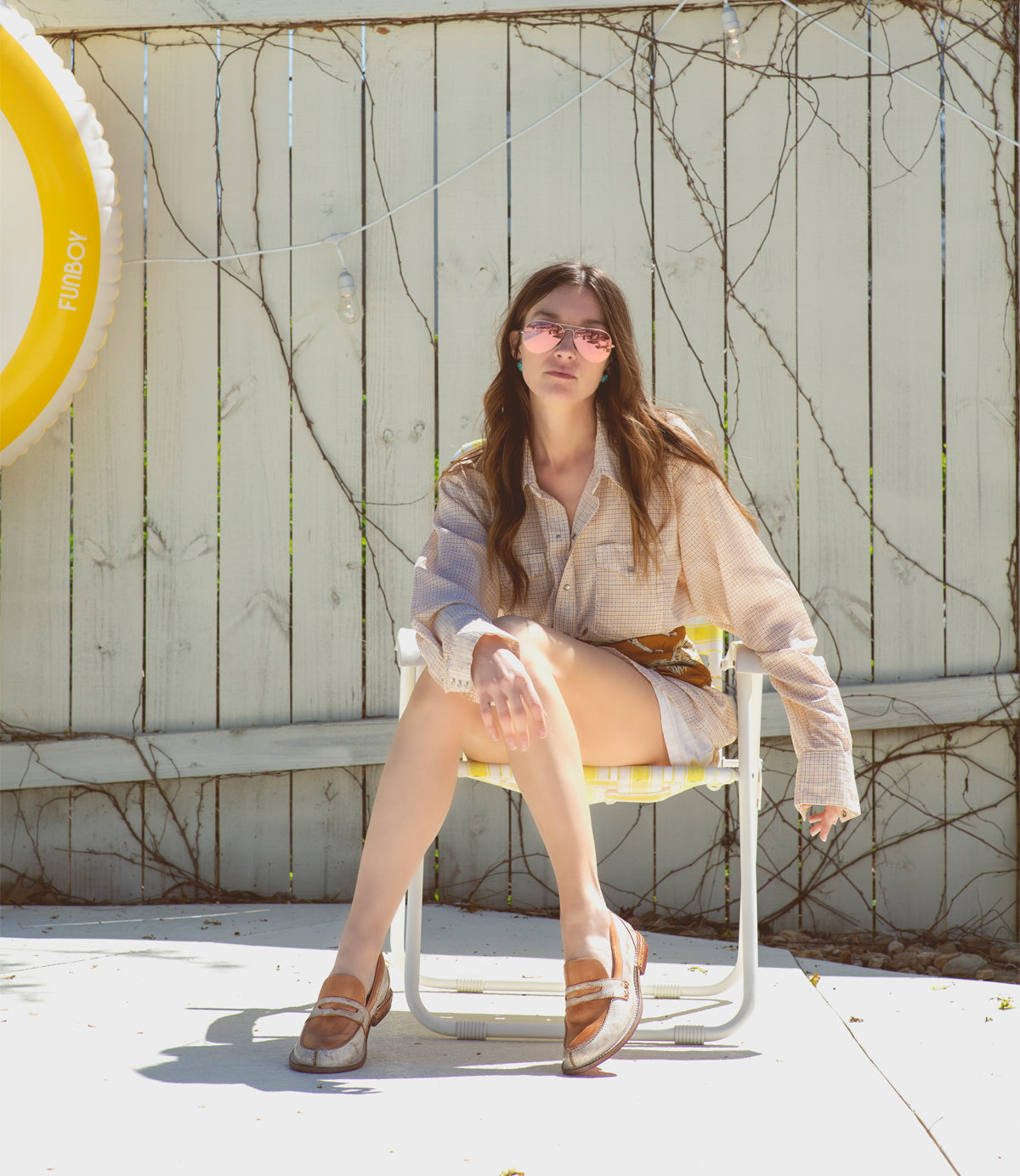 A woman wearing sunglasses and a beige outfit sits on a metal chair outside by a white fence draped with vines, her Bed|Stü Reina penny loafers—with their classic leather upper—resting casually at her side. A yellow pool float hangs on the fence.