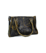 The Bed|Stü Rockababy is a dark brown distressed leather tote bag with dual handles, decorative side straps, visible stitching, and a weathered finish—crafted from vegetable-tanned leather—shown against a white background.