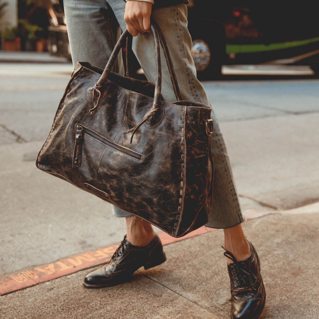 A person stands on a sidewalk near the street, holding the Rockaway, a large, vegetable-tanned leather handbag by Bed|Stü, paired with cuffed jeans and black oxford shoes.