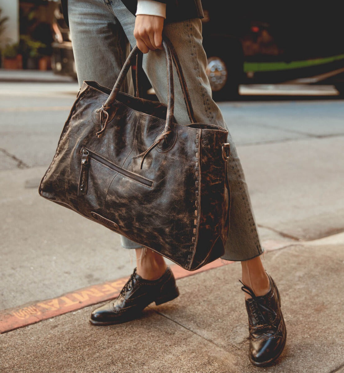 A person stands on a sidewalk near the street, holding the Rockaway, a large, vegetable-tanned leather handbag by Bed|Stü, paired with cuffed jeans and black oxford shoes.