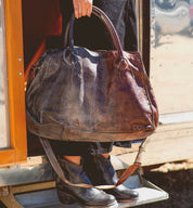 A person stands on a step next to an open door, wearing black pants and boots, while holding the Bed|Stü Rockaway—a large, worn full-grain leather bag featuring two handles and a shoulder strap.