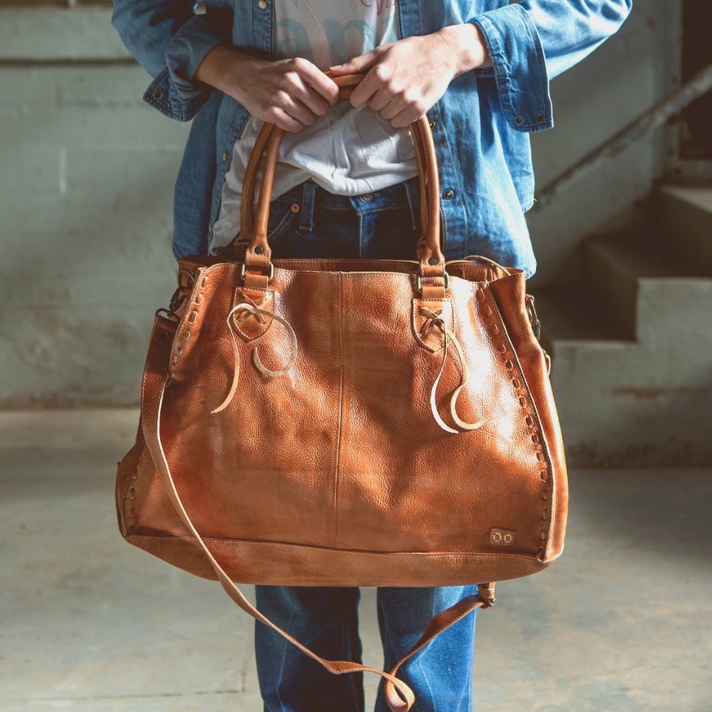 A person stands indoors wearing a denim jacket and jeans, holding the Bed|Stü Rockaway large brown vegetable-tanned leather handbag by its handles.