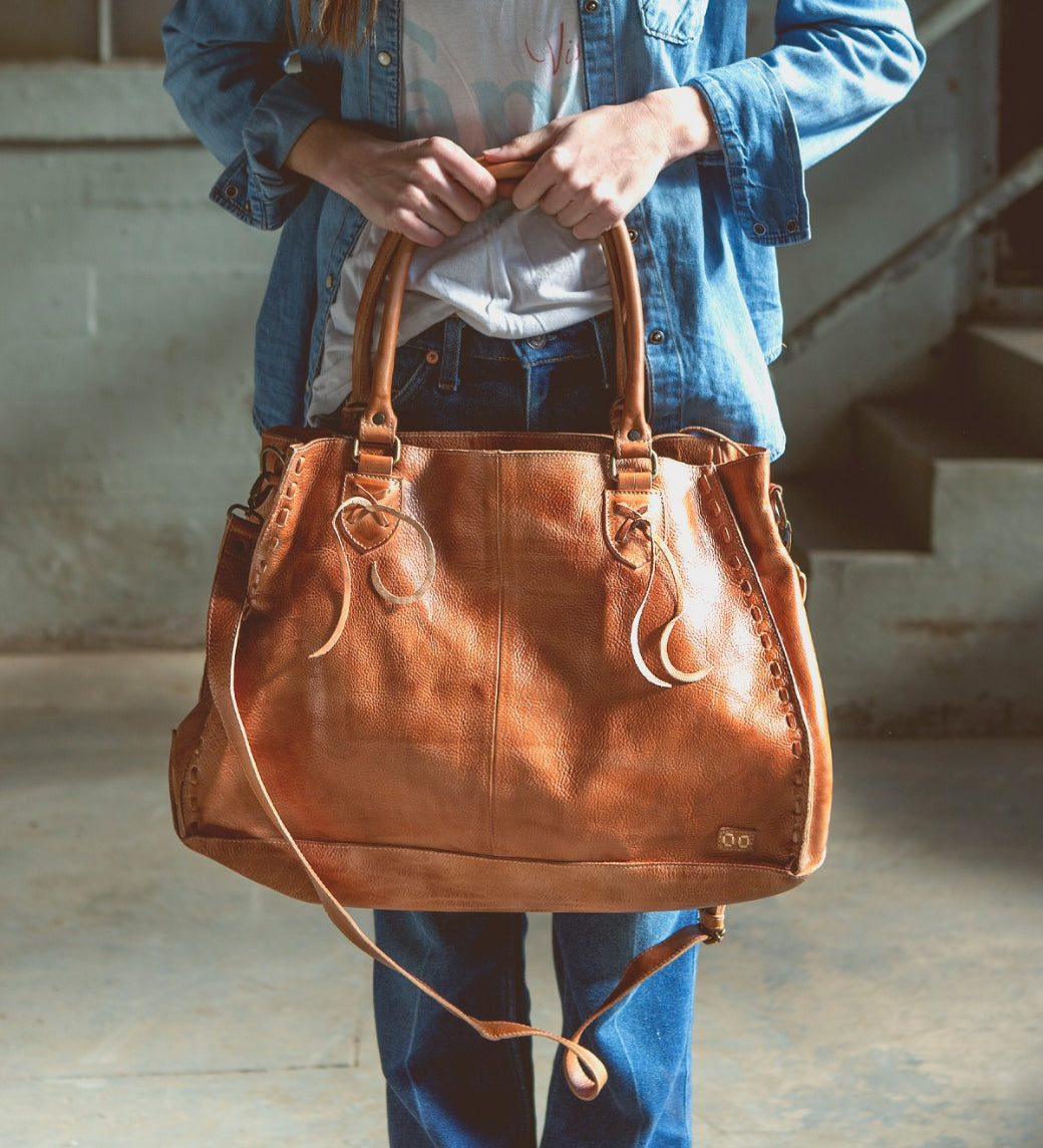 A person stands indoors wearing a denim jacket and jeans, holding the Bed|Stü Rockaway large brown vegetable-tanned leather handbag by its handles.