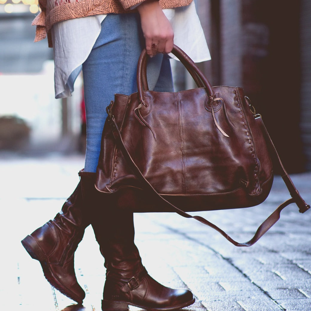 A person wearing blue jeans, brown boots, and a patterned jacket stands on a wet brick path, holding the Rockaway by Bed|Stü—a large handbag crafted from vegetable-tanned leather—by its handles: a timeless accessory that blends everyday function with classic style.