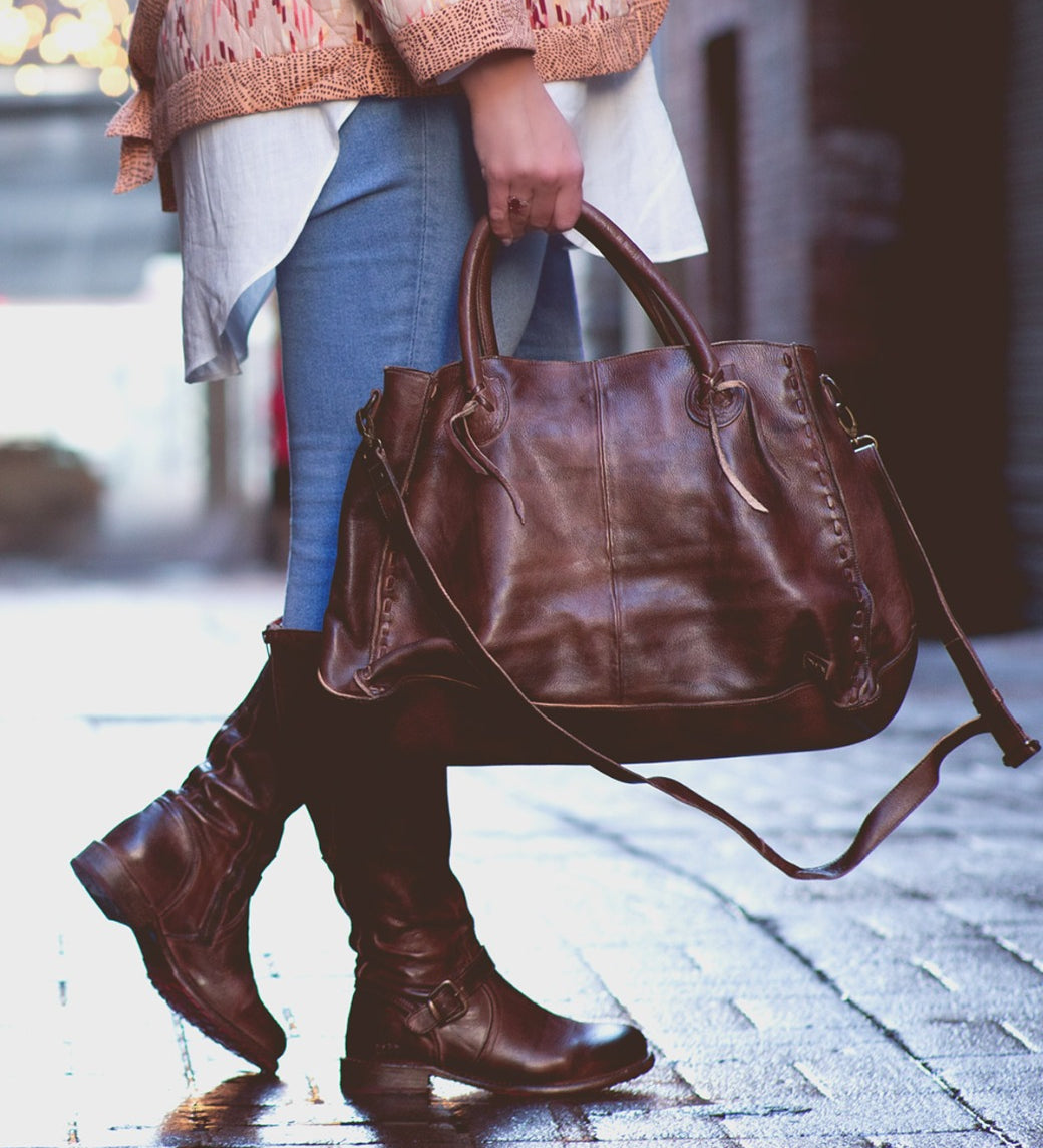 A person wearing blue jeans, brown boots, and a patterned jacket stands on a wet brick path, holding the Rockaway by Bed|Stü—a large handbag crafted from vegetable-tanned leather—by its handles: a timeless accessory that blends everyday function with classic style.