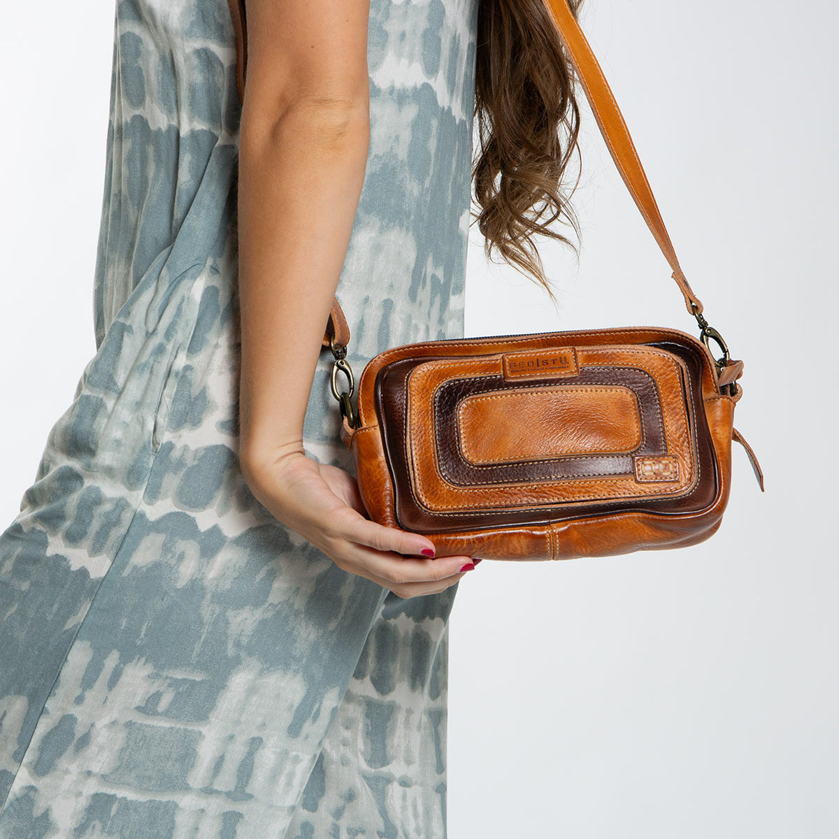 A woman in a blue and white dress carries the Bed|Stü Shy, a compact leather bag with dual pockets, featuring a small brown crossbody design and a convenient front pocket.