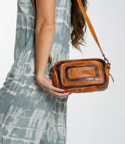 A woman in a blue and white dress carries the Bed|Stü Shy, a compact leather bag with dual pockets, featuring a small brown crossbody design and a convenient front pocket.