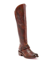 The Bed|Stü Takoma is a tall brown leather riding boot with a low heel, rounded toe, and braided detail across the ankle. Finished with an asymmetrical top for added style, this boot is pictured upright against a white background.