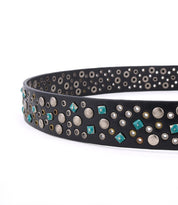 The Tammin II belt by Bed|Stü is a black leather accessory with a silver buckle, adorned with an eclectic mix of silver, gold, and turquoise studs in assorted shapes and sizes arranged in a random pattern.