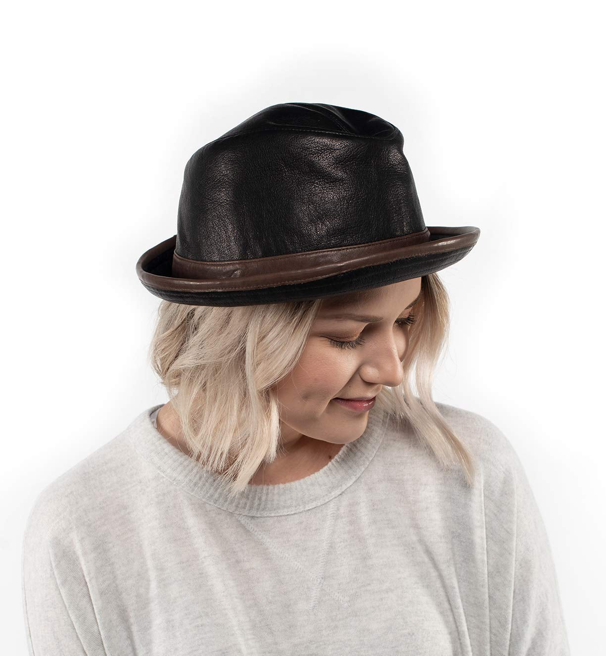 A person with blonde hair wears the Turino, a lightweight, handmade leather hat by Bed|Stü featuring a brown brim. They are looking down and smiling, dressed in a light-colored top against a plain white background.