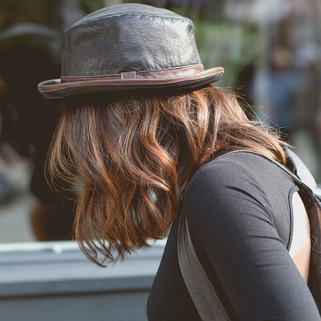 A person with wavy brown hair, wearing the Bed|Stü Turino lightweight black hat and a black top, is facing away from the camera outdoors.