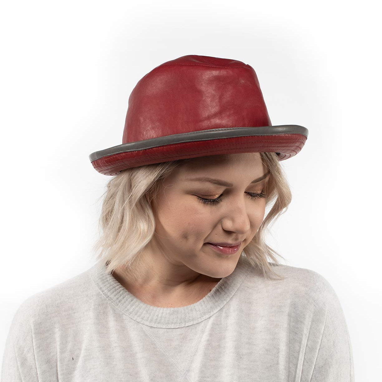 A woman with blonde hair wears the handmade Turino red leather hat by Bed|Stü and a light gray sweater, looking down with a neutral expression against a white background. The Turino’s lightweight, crushable design adds both comfort and style to her look.