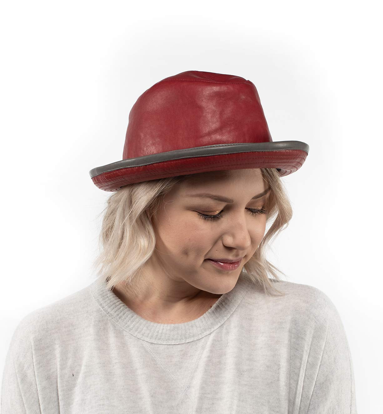 A woman with blonde hair wears the handmade Turino red leather hat by Bed|Stü and a light gray sweater, looking down with a neutral expression against a white background. The Turino’s lightweight, crushable design adds both comfort and style to her look.