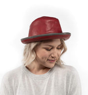 A woman with blonde hair wears the handmade Turino red leather hat by Bed|Stü and a light gray sweater, looking down with a neutral expression against a white background. The Turino’s lightweight, crushable design adds both comfort and style to her look.