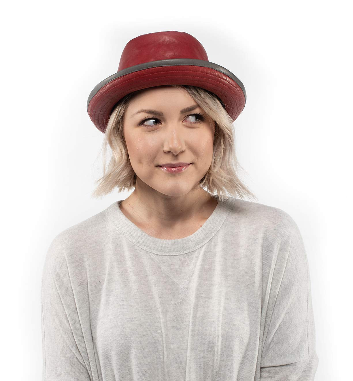 A woman with short blonde hair wears the Bed|Stü Turino crushable, handmade leather hat and a light gray sweater, looking to the side with a neutral expression against a white background.
