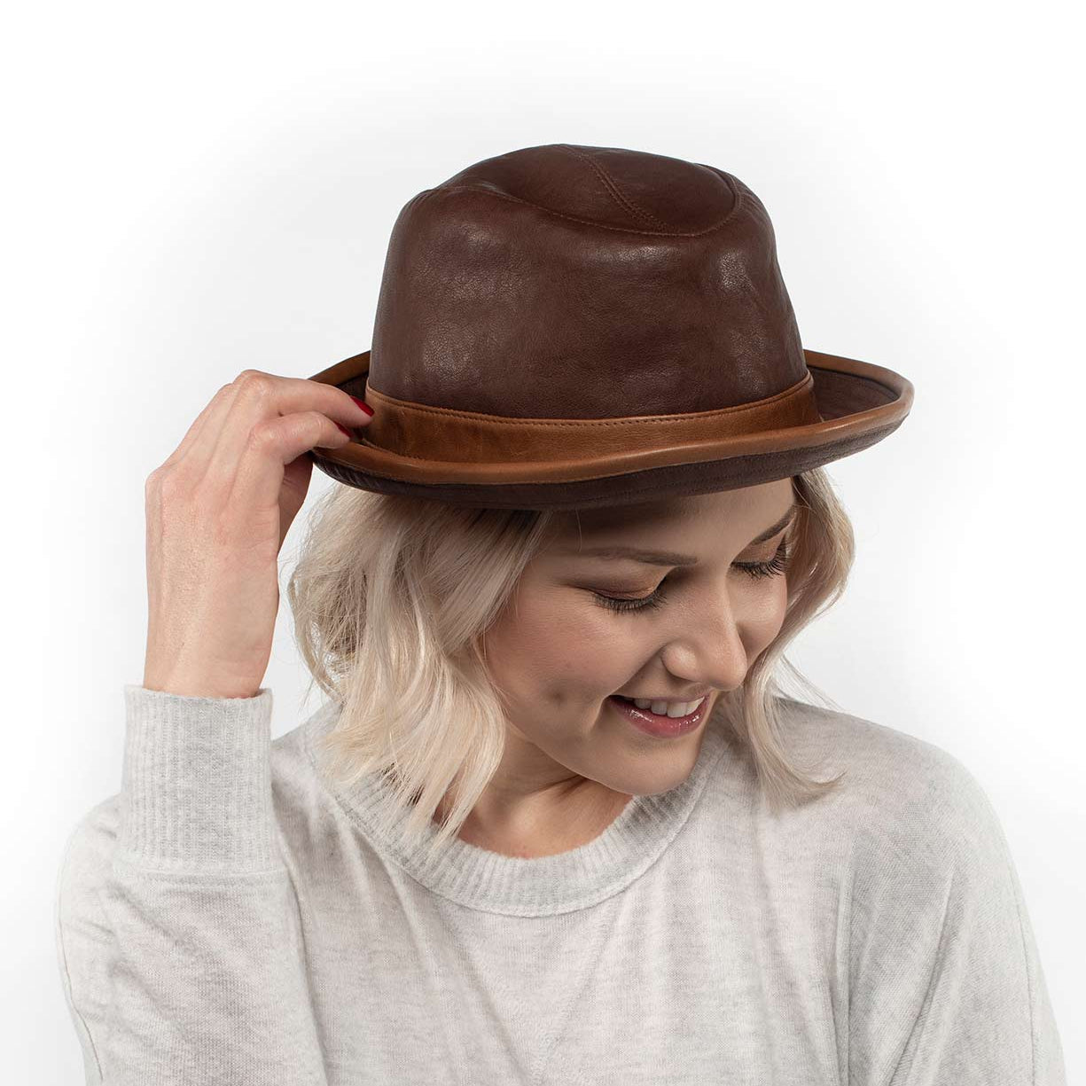 A person with light blonde hair, dressed in a light sweater, smiles as they adjust the Turino crushable handmade leather hat by Bed|Stü while looking down.