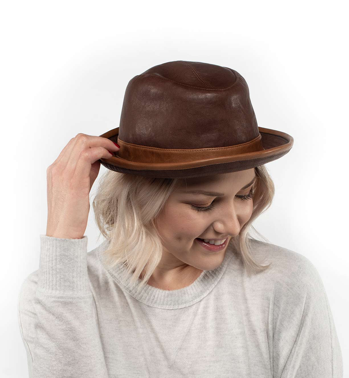 A person with light blonde hair, dressed in a light sweater, smiles as they adjust the Turino crushable handmade leather hat by Bed|Stü while looking down.