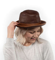A person with light blonde hair, dressed in a light sweater, smiles as they adjust the Turino crushable handmade leather hat by Bed|Stü while looking down.