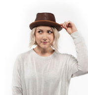 A person with short blonde hair wears the Turino, a brown crushable handmade leather hat by Bed|Stü, paired with a light sweater. They hold the brim of the hat with one hand and gaze slightly to the side against a white background.