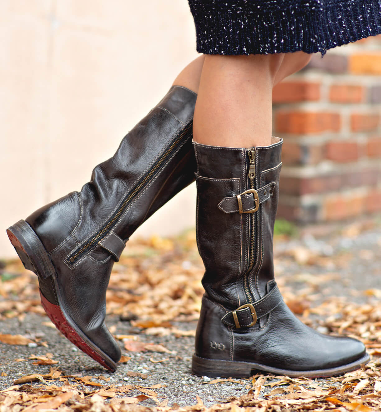 A person wearing Bed|Stü Gogo Lug Wide Calf black leather engineer boots with buckles and zippers stands on a leaf-strewn path near a brick wall.