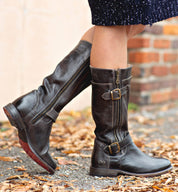 A person wearing Bed|Stü Gogo Lug Wide Calf black leather engineer boots with buckles and zippers stands on a leaf-strewn path near a brick wall.