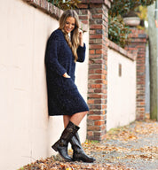 A woman wearing the Bed|Stü Gogo Lug Wide Calf black leather boots pairs them with a navy blue sweater dress as she stands outdoors against a brick wall, smiling and looking down.