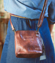 A person carrying the Bed|Stü Ventura, a brown leather crossbody bag featuring embossed text and an exterior zip pocket, while wearing a blue dress and standing outdoors.