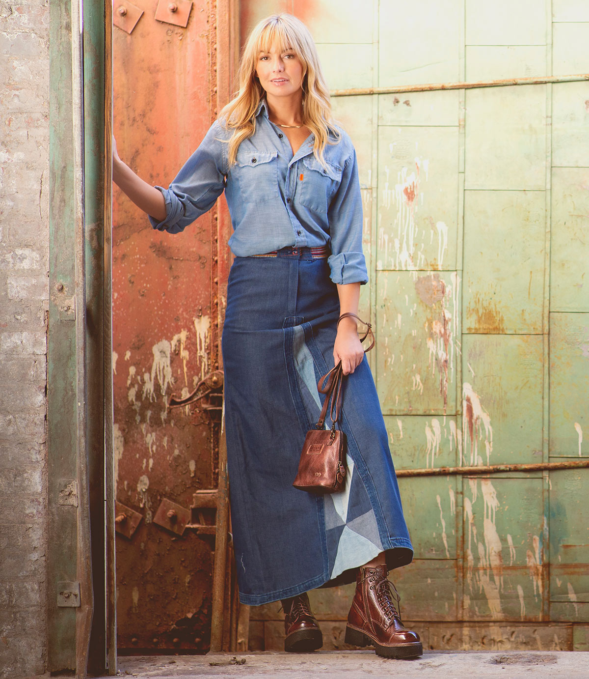 A woman stands in front of a rustic, industrial doorway wearing a denim shirt, long denim skirt, brown belt, and brown boots, while holding the Ventura lightweight purse with an exterior zip pocket by Bed|Stü.