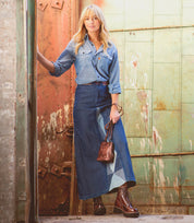 A woman stands in front of a rustic, industrial doorway wearing a denim shirt, long denim skirt, brown belt, and brown boots, while holding the Ventura lightweight purse with an exterior zip pocket by Bed|Stü.